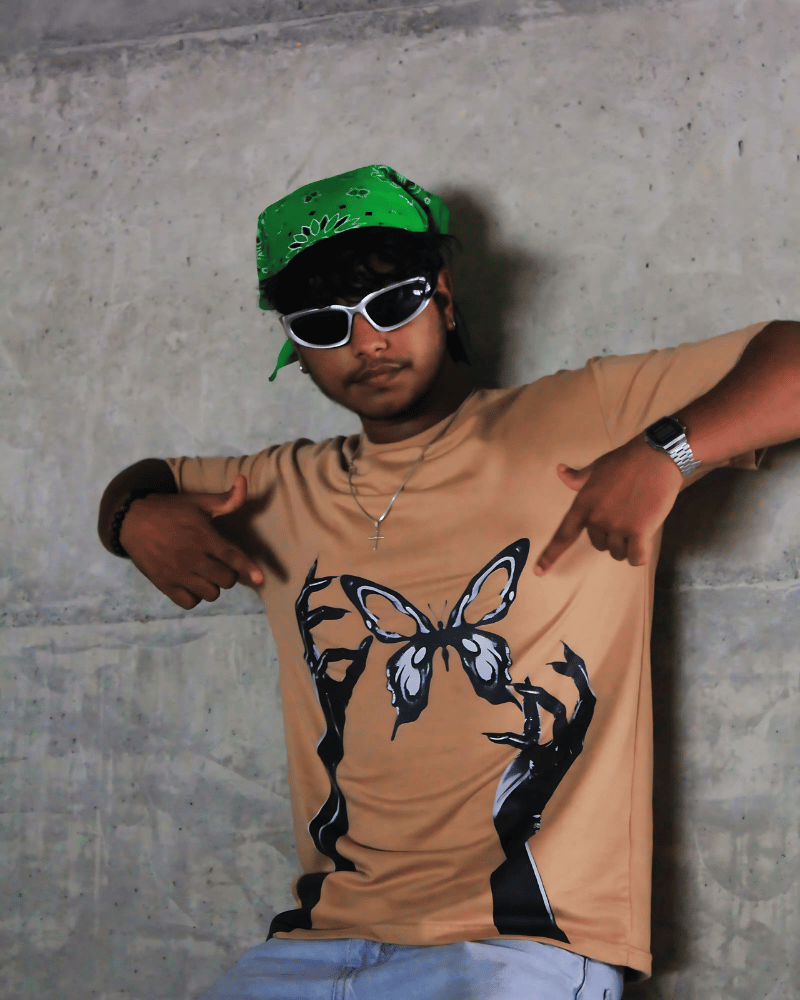Man wearing tan tainted flight graphic t-shirt with black hand and butterfly design posing against concrete wall